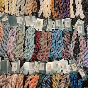 Puede incluir: Una colección de madejas de hilo de bordar de colores en varios tonos, incluyendo azules, verdes, rosas y marrones. Cada madeja está cuidadosamente organizada con una etiqueta adjunta, que muestra la marca y la información del color. El hilo parece estar hecho de un material suave y de alta calidad.