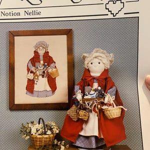 May include: A cross-stitch pattern for a doll called "Notion Nellie" from the HeartStrings collection. The doll is dressed in a red cape and a blue and white dress. She is carrying a basket of items, including a teapot, a cup, and a spool of thread.