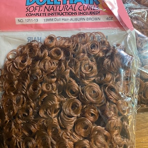 Puede incluir: Un paquete de pelo de muñeca marrón rojizo con rizos suaves y naturales. El paquete está etiquetado como "Doll Hair" e incluye instrucciones completas. El pelo mide 13 mm.