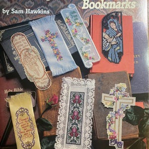 May include: A collection of cross-stitch bookmarks, including a cross, floral designs, and the word "LOVE". The cover of the book reads "Inspirational Bookmarks" by Sam Hawkins. The bookmarks are on top of old books.