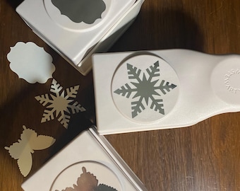 May include: Three white paper punches with different shapes: a label, a snowflake, and a butterfly. The punches are on a wooden surface, with cut-out shapes next to each punch. The punches are for crafting and scrapbooking.