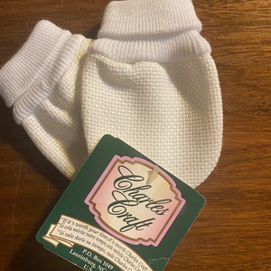 May include: A pair of white baby mittens with a folded cuff. The mittens are made of a soft, textured fabric. The tag attached to the mittens reads "Charles Craft" and includes the company's address and slogan: "If it's worth your time, it's worth Charles Craft."
