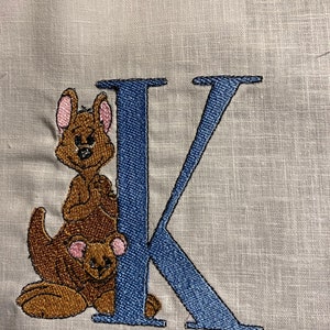 May include: Embroidered design of a blue letter K with two brown kangaroos peeking out from behind it. The kangaroos have pink ears and brown fur.