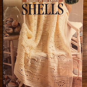 May include: A book cover for "Afghans by the Dozen SHELLS" showing a cream-coloured crocheted afghan draped over a chair. The cover includes the text "NEW" and "By Anne Halliday". The background includes seashells and a wicker basket.