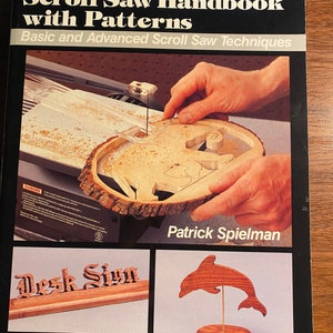 Peut inclure: Un livre intitulé "Scroll Saw Handbook with Patterns" de Patrick Spielman. La couverture du livre présente un gros plan d'une main utilisant une scie à chantourner pour découper un motif en bois. La couverture du livre comprend également des images d'une enseigne en bois et d'un dauphin en bois.