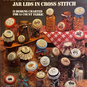 May include: A book cover titled "Canned Country" featuring various glass jars filled with different items, each topped with cross-stitched jar lids. The lids display charming designs and text. The cover also includes the text "21 Designs Charted for 14 Count Fabric."