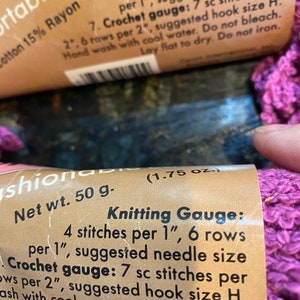 May include: Close-up of a pink yarn ball with a brown label that reads "Net wt. 50 g. (1.75 oz.) Knitting Gauge: 4 stitches per 1", 6 rows per 1", suggested needle size Crochet gauge: 7 sc stitches per 2", suggested hook size H. Wash with cool water. Do not bleach. Lay flat to dry. Do not iron."