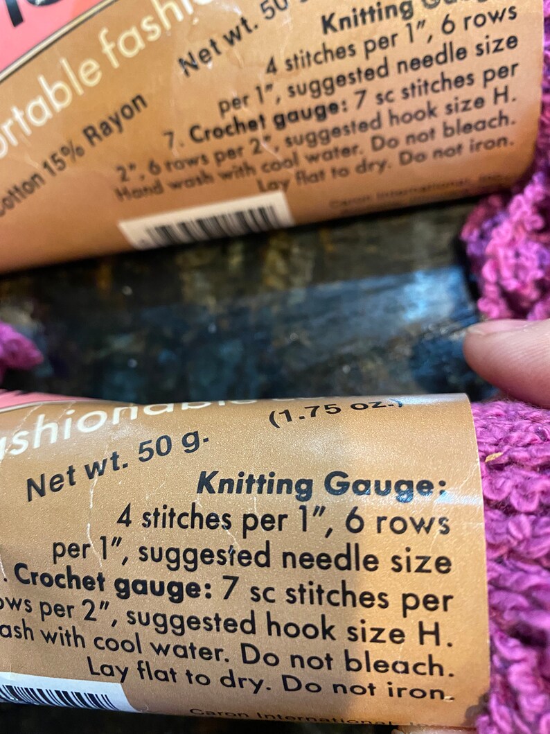 May include: Close-up of a pink yarn ball with a brown label that reads "Net wt. 50 g. (1.75 oz.) Knitting Gauge: 4 stitches per 1", 6 rows per 1", suggested needle size Crochet gauge: 7 sc stitches per 2", suggested hook size H. Wash with cool water. Do not bleach. Lay flat to dry. Do not iron."