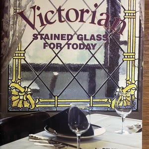 May include: A book cover titled Victorian Stained Glass for Today by Bill Hillman, featuring a graphic of a leaded glass window with yellow accents above a white-clothed dining table set with wine glasses, a dark napkin, and silverware.