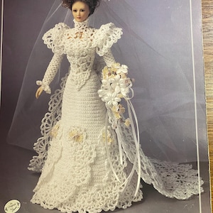 May include: A white crochet doll in a wedding dress, complete with a veil and bouquet. The dress has long sleeves, a high collar, and a detailed lace design. The doll has brown hair and is set against a grey backdrop.
