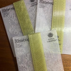 May include: A collection of Cricut Cuttlebug embossing folders and matching yellow spacers. The folders feature intricate lace and floral designs. The Anna Griffin brand is visible on the folders. The items are arranged on a wooden surface.