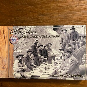 Op de afbeelding: Een vintage zwart-witfoto van een groep mannen die buiten aan een tafel zitten. De foto staat op de omslag van een boek getiteld "White Pass Recipe Card Collection" met 24 recepten. Het boek heeft een rood-wit logo met de woorden "White Pass & Yukon Route".