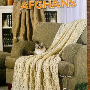 May include: A crochet book cover titled "Timeless, Textured AFGHANS" featuring a cream-colored afghan draped over a green armchair. The afghan has a cable knit design. A cat is resting on the chair. The book cover also shows a second afghan.