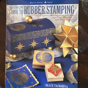 May include: The Complete Guide to Rubber Stamping book cover features a blue background with gold stars and a gold star-shaped ornament. The book title is in white with a gold outline. The book cover also features a wooden box with a gold moon and stars design, and three rubber stamps with a moon, star, and a crescent moon design.