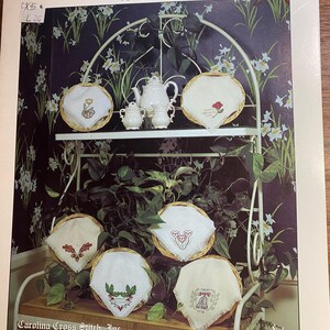 Op de afbeelding: Een display van geborduurde broodhoezen in geweven manden, gerangschikt op een wit metalen rek. De hoezen hebben bloemen en decoratieve ontwerpen. Een witte theepot en suikerpot set zijn ook aanwezig. De tekst "Special Occasions Breadcovers" is zichtbaar.