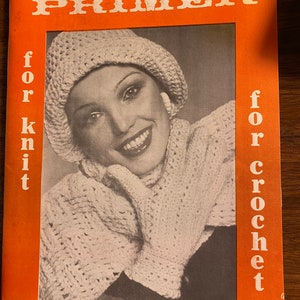 Può includere: La copertina di The Needlework Primer presenta una donna con un cappello e un maglione di uncinetto bianco. La copertina è arancione con il testo "The Needlework Primer for knit for crochet" in bianco.