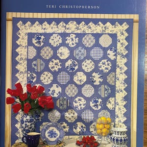 May include: A book cover titled "Days to Remember" by Teri Christopherson. The cover features a blue and white quilt design with floral patterns. A vase of red tulips, a blue cup, and a plate are also visible.