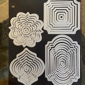 Puede incluir: Cuatro troqueles metálicos anidados en varias formas, incluyendo diseños de flores, cuadrados y arqueados, están dispuestos sobre una superficie negra. Cada troquel tiene un borde cosido, y las formas varían en tamaño, creando un efecto en capas.