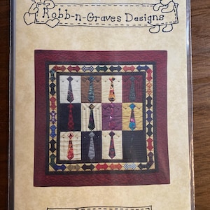 May include: A quilt pattern titled "Tie One On" featuring a patchwork design of ties on shirts. The quilt has a burgundy border and a decorative outer edge. The packaging includes the text "Robb-n-Graves Designs" and two bear illustrations.