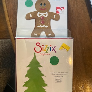 May include: Two Sizzix die-cut packages. The top package features a gingerbread man with a smiling face and a red button. The bottom package shows a green Christmas tree design. Both packages include a green circle.