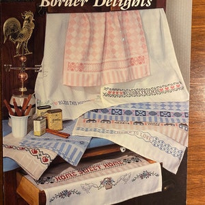 Può includere: Opuscolo "Border Delights" con schemi a punto croce per canovacci e decorazioni per la casa. I disegni includono motivi floreali, a cuore e geometrici, con frasi come "Home Sweet Home" e "Welcome Friends".