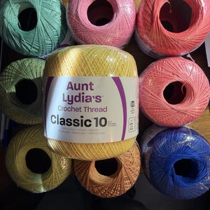 Op de afbeelding: Een verzameling van acht bollen Aunt Lydia's Classic 10 haakgaren in diverse kleuren, waaronder groen, roze, koraal, geel, beige en blauw. De middelste gele bol heeft een label met de tekst Aunt Lydia's Crochet Thread Classic 10.