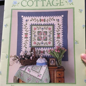 Op de afbeelding: Een groen en wit quiltpatroonboek getiteld "Rosewood Cottage" van Brandywine Design. De omslag toont een quilt met een bloemenontwerp in tinten roze, groen en wit. De quilt wordt getoond op een tafel met een pop en een vaas met tulpen.
