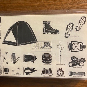 May include: A sheet of black and white camping-themed rubber stamps. Designs include a tent, hiking boot, footprints, a lantern, logs, binoculars, a compass, and other outdoor elements. The text "ROUGHING IT" is visible.