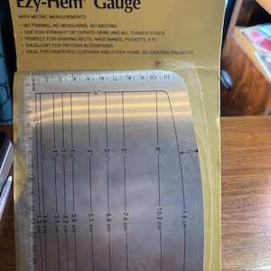 May include: A silver Ezy-Hem Gauge with metric measurements for sewing hems and shaping fabric. The gauge has markings in inches and centimeters up to 4 inches.