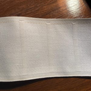 May include: Close-up of a section of white woven fabric with a textured, herringbone pattern. The fabric appears to be a sturdy material, possibly for upholstery or crafting. The edges are neatly finished with visible stitching.