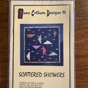 May include: A package for a quilt pattern titled "Scattered Showers" by June Colburn Designs 11. The pattern features a design of colorful umbrellas on a dark blue background. The text "Celebrate the rain by creating this fun, easy wall quilt with paper piecing and applique!" is also visible.