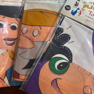 May include: Vintage sewing patterns for "Toytown" and "The Flintstones" dolls. The patterns feature cartoon character faces in bright colors. The packaging includes instructions and the "Springmaid Fabric" logo. The patterns are for sew-it-yourself dolls.