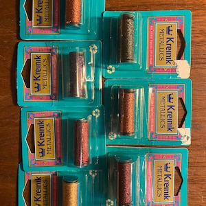 Può includere: Sette bobine di filo da ricamo metallizzato Kreinik in vari colori, tra cui oro, rame, argento e rosso. Ogni bobina è confezionata in un contenitore di plastica trasparente con un'etichetta blu e bianca che recita "Kreinik Metallics".