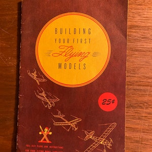 Pode incluir: Um livreto vermelho com um círculo amarelo na frente que diz "Building Your First Flying Models". O livreto tem um preço de 25 centavos e mostra ilustrações de aviões modelo.