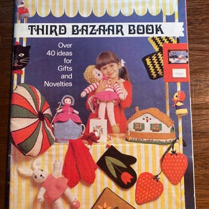 May include: A book cover with the title "Third Bazaar Book" and a bee logo. The cover features a variety of knitted items, including a doll, a house, a pair of mittens, and a strawberry. The book is from Patons, a yarn company.