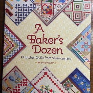 Pode incluir: Um livro intitulado "A Baker's Dozen" com 13 colchas de cozinha de American Jane. A capa é amarelo claro com um padrão de pequenos círculos e apresenta vários designs de colchas nos cantos, incluindo cestos, galinhas e padrões florais.