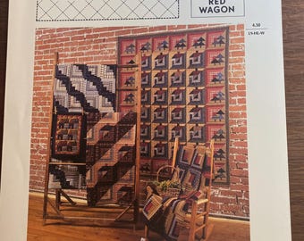 Op de afbeelding: Een boekomslag met de titel "HOUSES LOG CABINS" met een Red Wagon-logo. De afbeelding toont quilts met blokhutontwerpen die worden getoond op houten standaards en een schommelstoel. De quilts hebben verschillende kleuren en patronen, met een bakstenen muur als achtergrond.