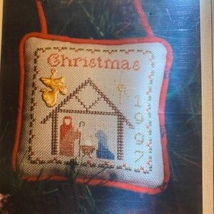 May include: A white and red Christmas ornament with a cross-stitch design depicting the nativity scene. The ornament says "Christmas 1997" and features a star, an angel, and a manger with the baby Jesus, Mary, and Joseph.