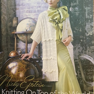 May include: A book cover titled Nicky Epstein Knitting On Top of the World The Global Guide to Traditions, Techniques and Design. It features a person wearing a cream-colored, open-front, tiered lace cardigan over a long, light green dress.