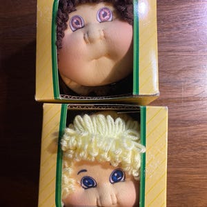 May include: Two Cabbage Patch Kids ear muffs in their original boxes. One has brown hair and the other has blonde hair. The boxes are yellow with green trim and the Cabbage Patch Kids logo.