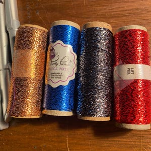 May include: Four spools of metallic thread in copper, blue, silver, and red. The blue spool has a label that reads "Pretty Fun Stuff Angel Hair Washable Colorfast 225 Yards". The red spool has a label that reads "Lift Peel".