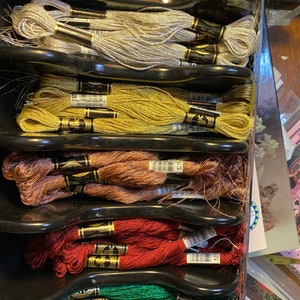 May include: A close-up of a black plastic organizer filled with spools of metallic embroidery floss. The floss is arranged in rows by color, starting with gold, silver, yellow, copper, red, and green.