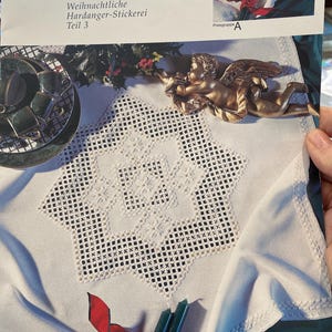 May include: A white tablecloth with a detailed cross-stitch design, featuring a central star pattern. The image also shows a gold-colored angel figurine, a set of green and black teacups, and the cover of a craft book titled "Intermezzo".