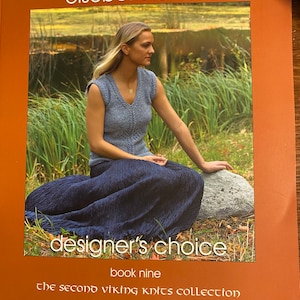 May include: A book cover featuring a woman wearing a blue knitted top and a long blue skirt, sitting on a rock in a grassy area. The book title is "designer's choice" and the subtitle is "the second viking knits collection".