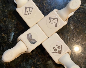 May include: Four cream-coloured corner paper punches with various decorative designs. Each punch features a handle and a corner design. The punches are arranged in a cross formation on a dark, speckled surface.