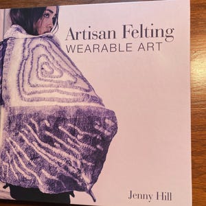 May include: A book titled "Artisan Felting: Wearable Art" by Jenny Hill. The cover features a close-up of a woman wearing a purple and white felted garment.