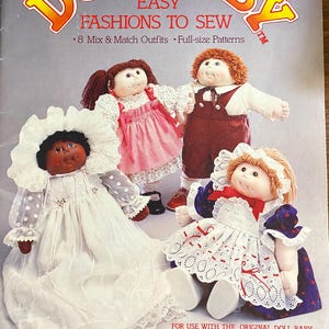 Puede incluir: Un libro de patrones titulado The Original Doll Baby Easy Fashions To Sew con 8 conjuntos combinables y patrones a tamaño real. La portada muestra tres muñecas de tela con vestidos y petos detallados con encajes y cintas.