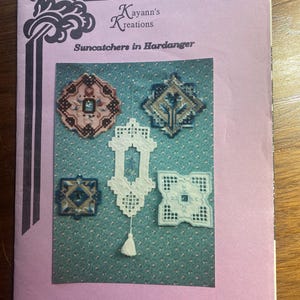 Op de afbeelding: Een roze brochure getiteld "Kayann's Kreations Suncatchers in Hardanger" toont verschillende handgemaakte zonnevangers. De zonnevangers hebben verschillende vormen, waaronder vierkanten en diamanten, en zijn gemaakt met wit, blauw en bruin garen.