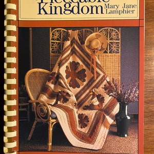 May include: A book titled "The Pieceable Kingdom" by Mary Jane Lampher, featuring a photograph of a quilt draped over a wicker chair with a straw hat on top of the quilt.