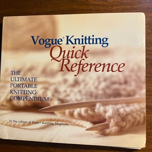 May include: A book titled "Vogue Knitting Quick Reference" with the subtitle "The Ultimate Portable Knitting Compendium." The cover features text in blue and red, with a blurred image of knitting needles and yarn. The book is by the editors of Vogue Knitting Magazine.